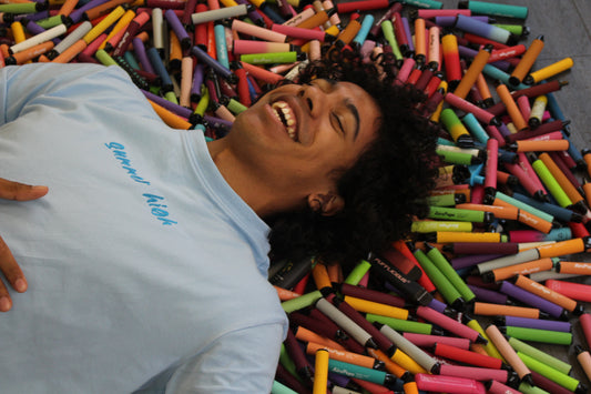 A man in a blue t-shirt with curly dark hair smiling while left hand on chest. Laying on a spread of colourful cylinder tubes.