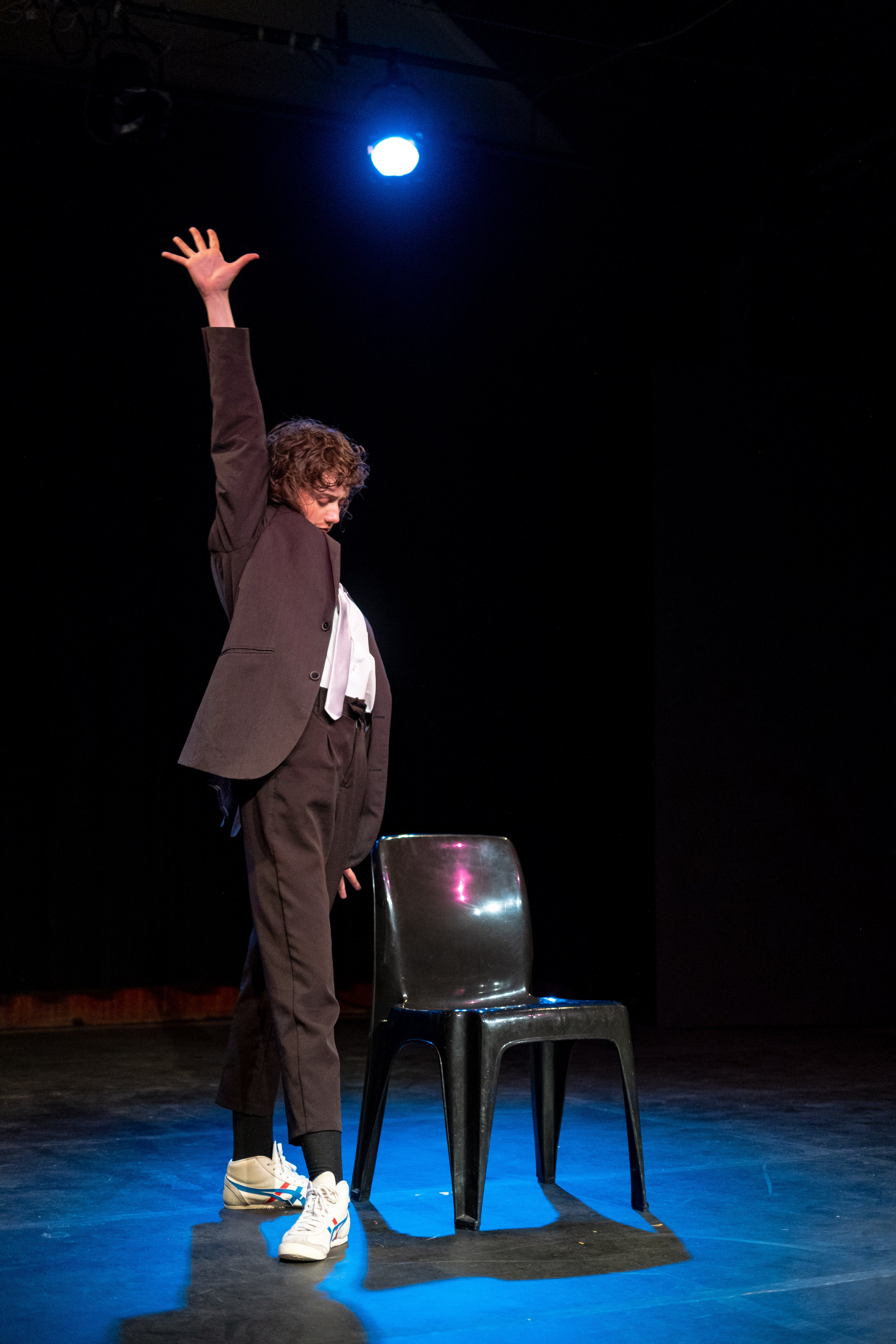 Person in a suit standing on a chair with one arm raised against a dark background